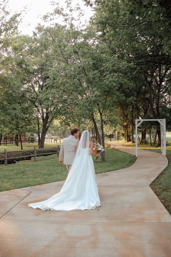 newlyweds walk on a tree-lined path through their outdoor wedding ceremony spot 