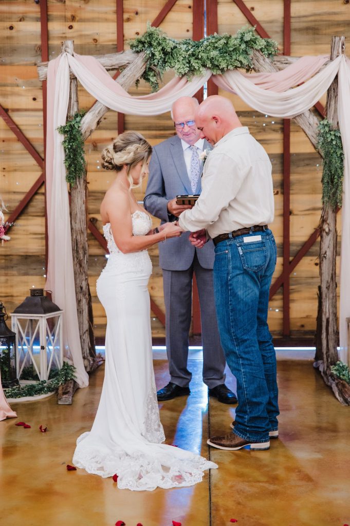 couple exchange rings at their indoor barn ceremony in front of a beautifully decorated wooden arch  